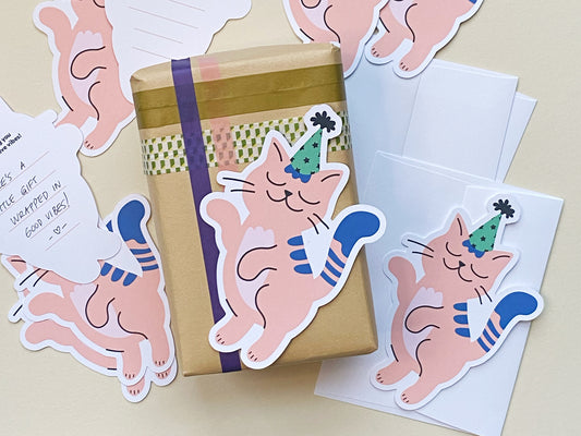 A small, gift-wrapped package with a kraft paper base and decorative washi tape. A pink cat die-cut note card is attached to the package. Surrounding the gift are several identical die-cut cards and white envelopes on a light background.