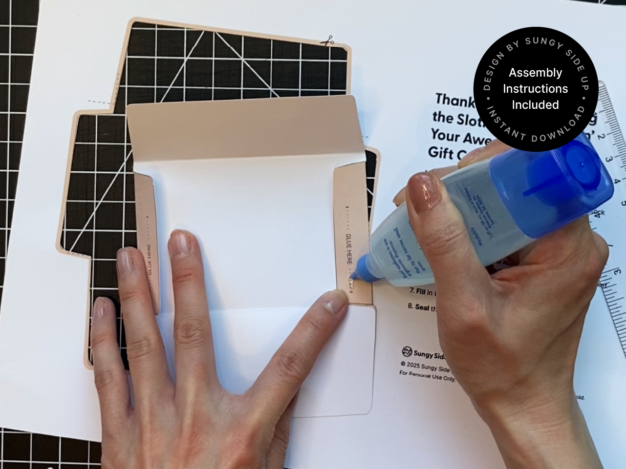 A pair of hands assembling the sloth gift card holder by applying blue liquid glue along the designated “Glue Here” tab. The template is printed on white paper with cut and fold lines visible, laid out on a black grid cutting mat for precision. The glue is applied carefully to ensure clean edges when the envelope is folded, demonstrating an easy and fun DIY crafting process for creating a personalized gift card holder.
