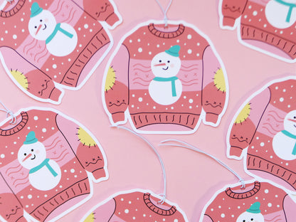 Several Christmas sweater-shaped gift tags, each with a pink background featuring a cheerful snowman with a teal hat and scarf. The sweaters have yellow elbow patches and snowflake designs, all set against a pink surface, adding to the festive and playful feel.
