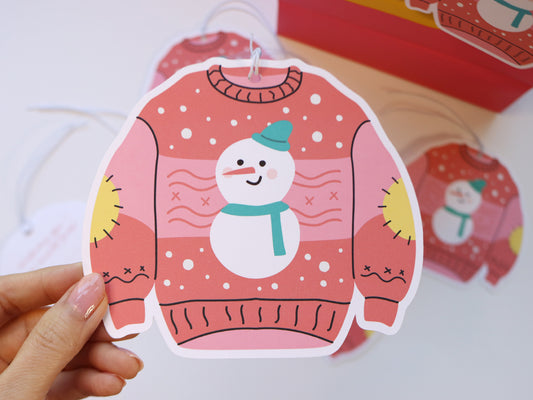 A close-up of a hand holding a pink Christmas sweater-shaped gift tag featuring a snowman with a teal hat and scarf. The festive design includes snowflakes and yellow elbow patches, perfect for adding cheer to holiday presents.