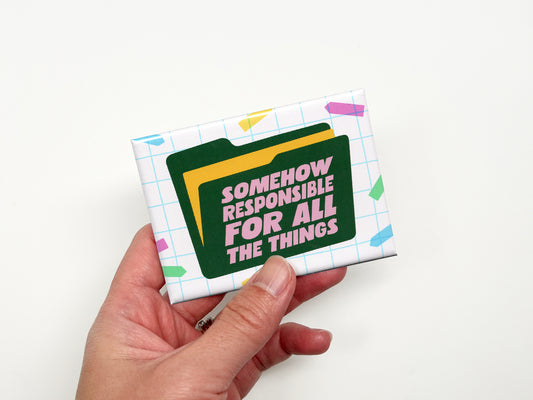 Hand holding a rectangular magnet with a white grid background and colorful confetti accents. A dark green file folder graphic features pink text that reads “Somehow Responsible for All the Things.”