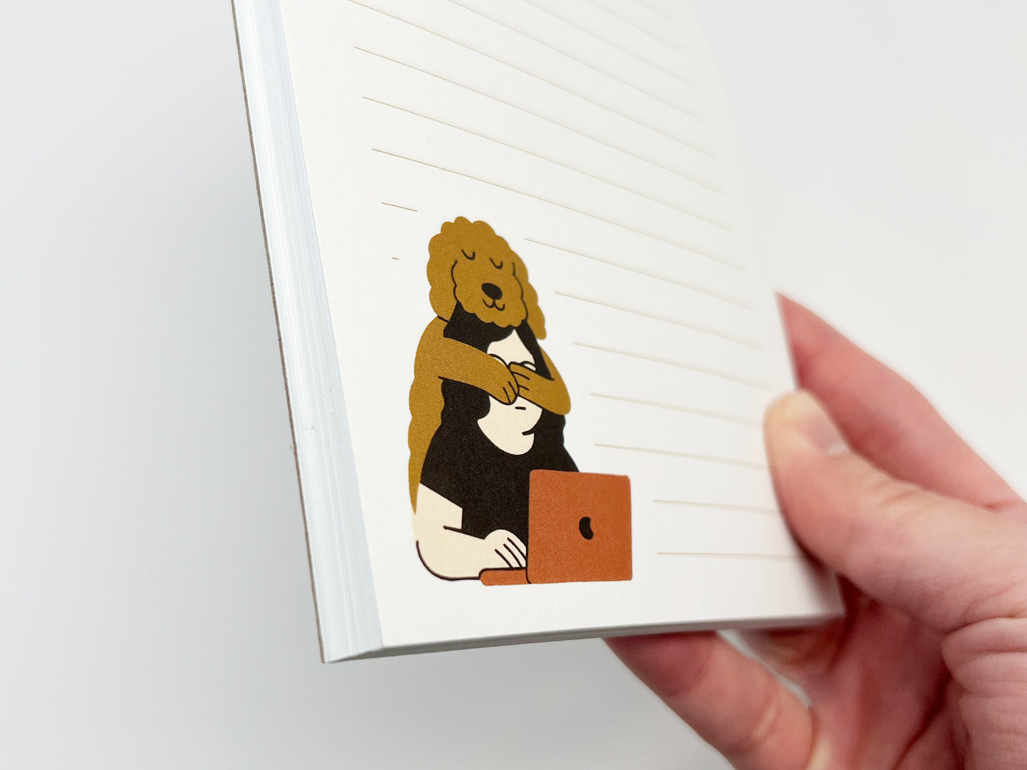 Angled close-up of a small lined notepad held in a hand, showing an illustration of a Golden Doodle hugging a person from behind as they work on a laptop, printed in the lower corner of the page.