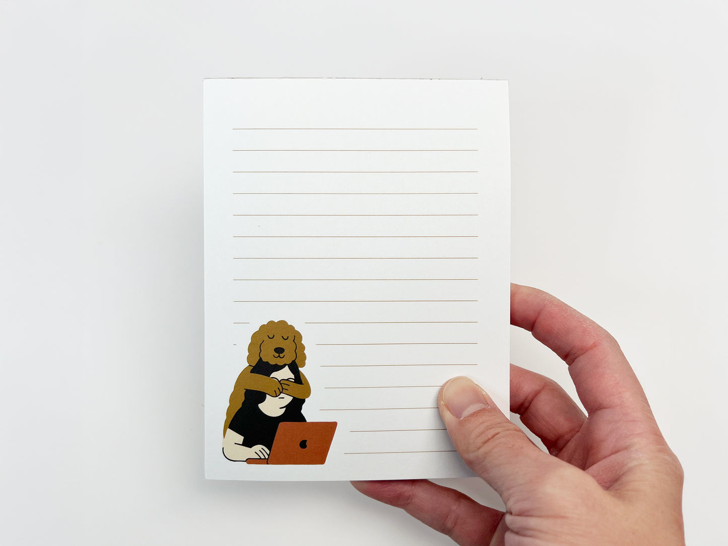 A hand holding a small lined notepad with white pages and light tan lines, featuring an illustration of a Golden Doodle resting its paws on a person working on a laptop in the lower left corner.