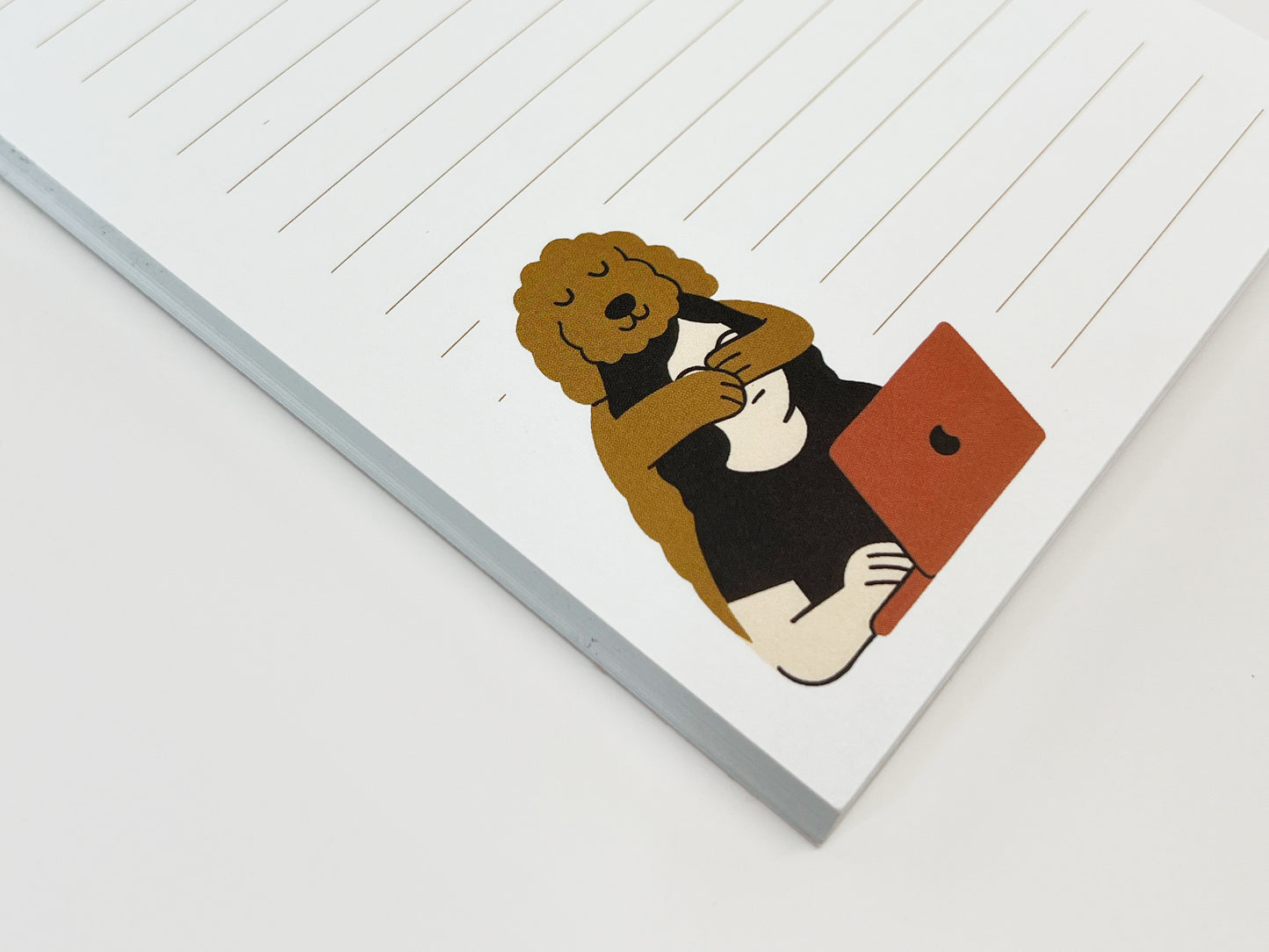 Angled flat lay close-up of a small lined notepad showing an illustrated Golden Doodle hugging a person who is working on a laptop, printed on white paper with light tan lines.