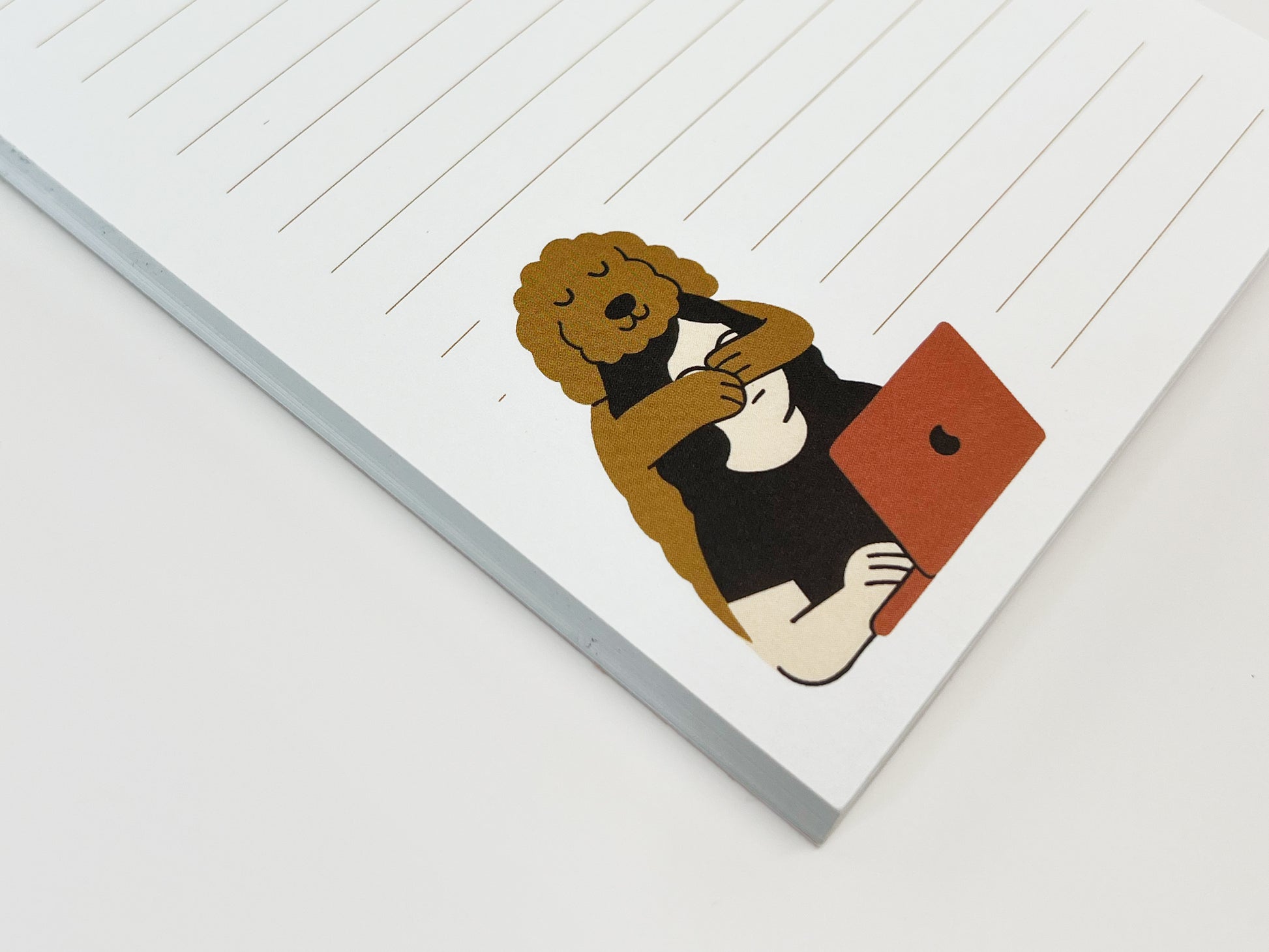 Angled flat lay close-up of a small lined notepad showing an illustrated Golden Doodle hugging a person who is working on a laptop, printed on white paper with light tan lines.