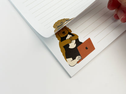Close-up of a lined notepad as a page is being lifted, revealing an illustration of a Golden Doodle hugging a person working on a laptop, printed in the lower corner of the page.