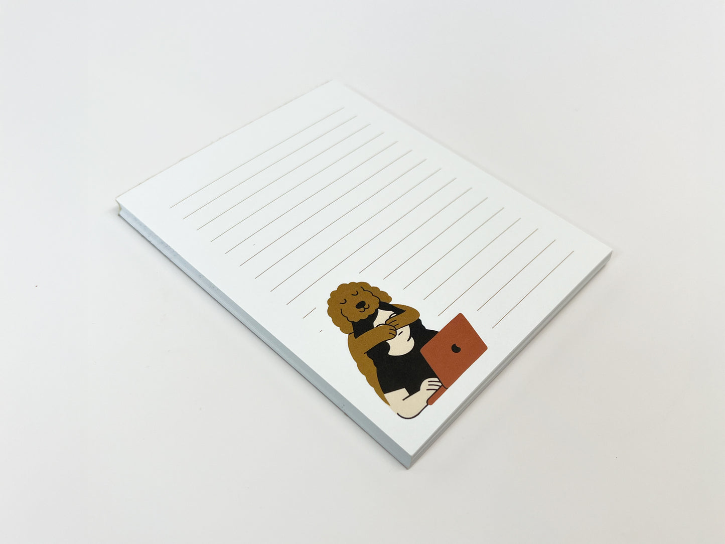 Angled flat lay of a small lined notepad with white pages and light tan lines, featuring an illustrated Golden Doodle hugging a person working on a laptop in the lower corner of the page.