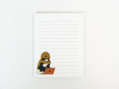 Top-down view of a small lined notepad with white pages and light tan lines, featuring an illustrated Golden Doodle hugging a person working on a laptop in the lower left corner.