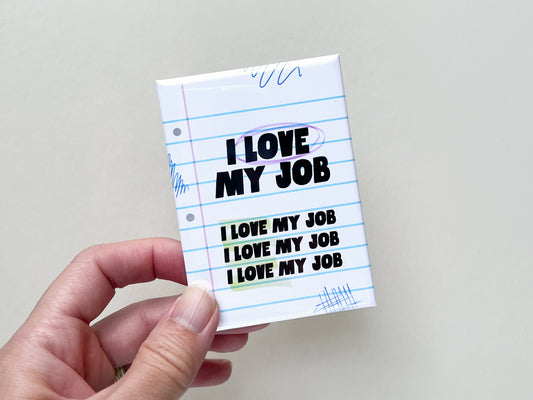 Hand holding a small rectangular fridge magnet with a notebook paper design. The magnet features bold black text reading “I LOVE MY JOB” at the top, with the phrase repeated three times below in smaller text. The background resembles lined paper with blue horizontal lines, a pink margin line, and small doodle scribbles in blue ink. The magnet has a glossy finish and rounded edges, shown against a plain light background.