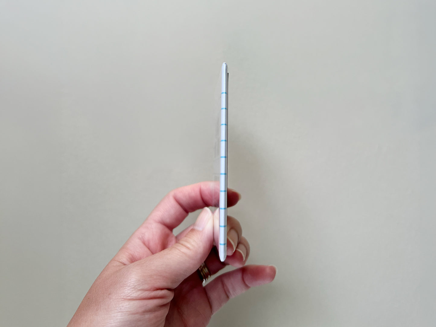 Side view of a small rectangular fridge magnet held between fingers, showing its slim thickness and slightly rounded edges. The magnet has a glossy finish with a notebook-style design visible along the edge, including light blue horizontal lines. The background is plain and light, keeping focus on the product’s thin profile.