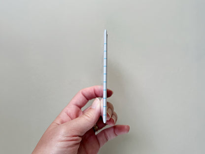 Side view of a small rectangular fridge magnet held between fingers, showing its slim thickness and slightly rounded edges. The magnet has a glossy finish with a notebook-style design visible along the edge, including light blue horizontal lines. The background is plain and light, keeping focus on the product’s thin profile.