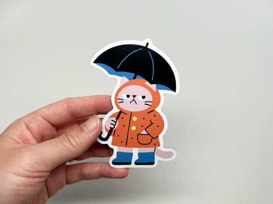 Hand holding a handmade die-cut magnet shaped like a grumpy cat in an orange raincoat with black umbrella, cute weather-themed gift for cat lovers and unique kitchen decor.
