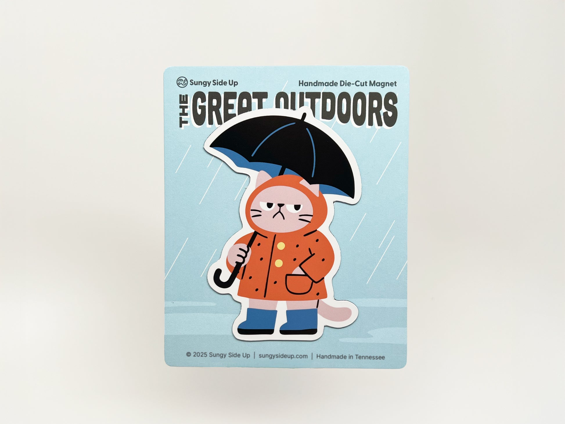 Handmade die-cut magnet of a grumpy cat wearing an orange raincoat and holding a black umbrella, displayed on blue illustrated backing card with raindrops, quirky weather-themed gift for cat lovers.