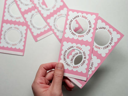 Close-up of the edge of an unassembled pink and white Valentines Day lollipop holder showing the adhesive strip for easy assembly. The design features scalloped edges, a circular cutout, and the text, XOXO You're Sweet Happy Valentine's Day!