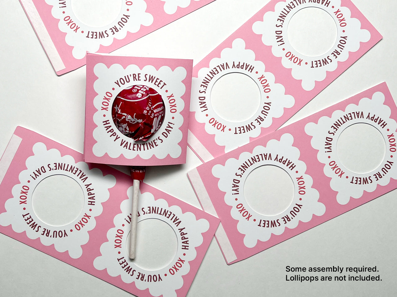 A Valentines Day lollipop holder with a round lollipop with a red wrapper inserted. Several unassembled pink and white holders are scattered around. These are ideal for kids classroom exchanges and party favors.