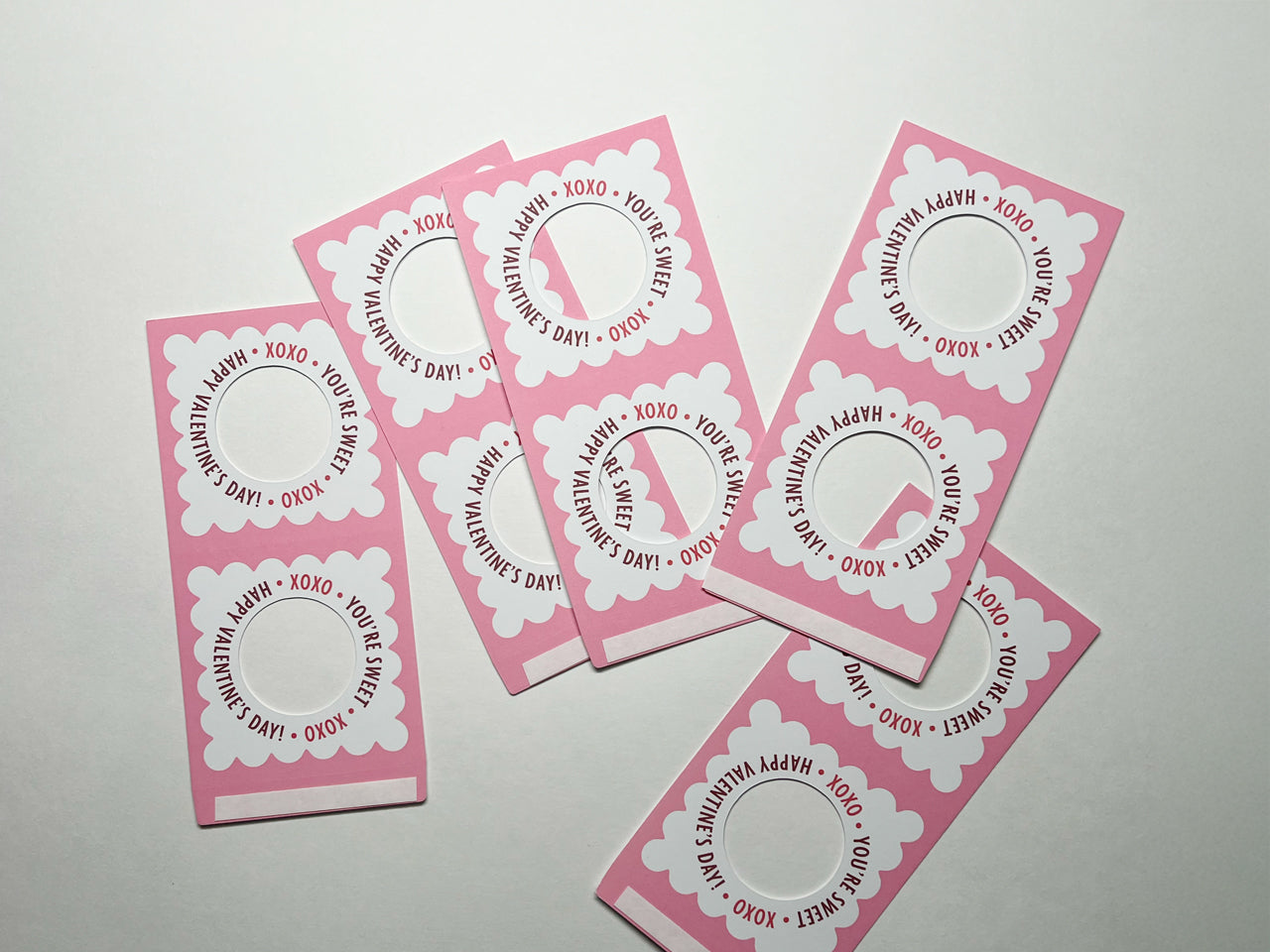 A group of pink and white lollipop holders for Valentines Day spread out on a white surface. Each lollipop holder has a scalloped design with the text, XOXO You're Sweet Happy Valentines Day! and a circular cutout for lollipops. Perfect for friends.