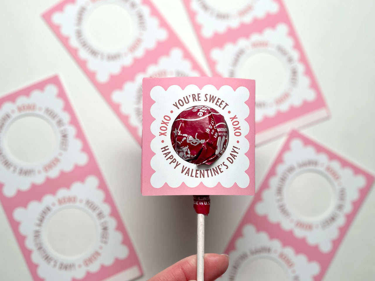 A close-up of a hand holding a pink and white Valentines Day lollipop holder with a round lollipop with red wrapper is inserted through the circular cutout. Several unassembled holders are blurred in the background.