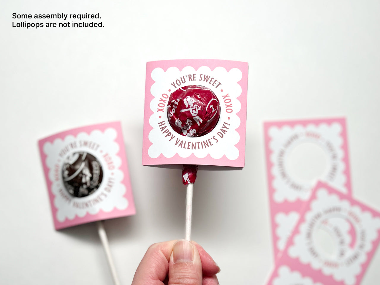 A hand holding a completed pink and white Valentines Day lollipop holder with a red lollipop inserted. Another lollipop with a brown wrapper is also lying in the background. Perfect for classroom exchanges and party favors.