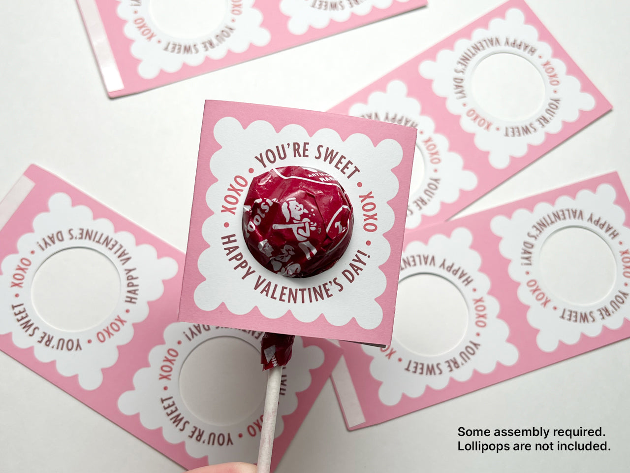 A close-up of a completed Valentines Day lollipop holder with a round lollipop with a red wrapper. The scalloped pink design says, XOXO You're Sweet Happy Valentines Day! The holder is surrounded by unassembled pieces.