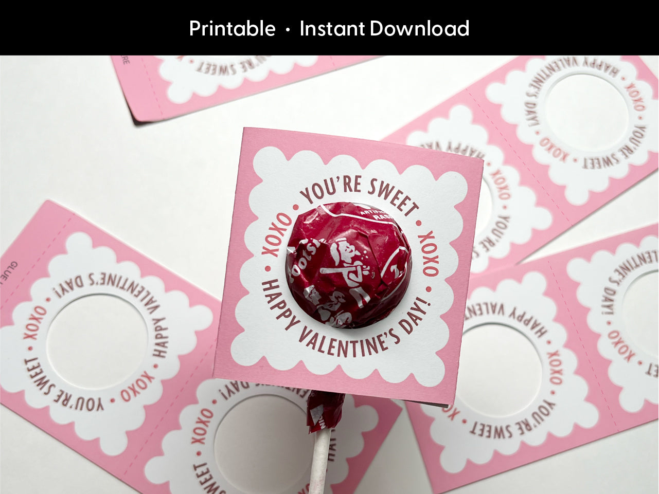 Assembled pink Valentines Day lollipop holder showcasing a scalloped edge design with a lollipop inserted. Surrounded by unassembled templates and printables, ideal for crafting Valentine gifts and party favors for classroom exchanges.