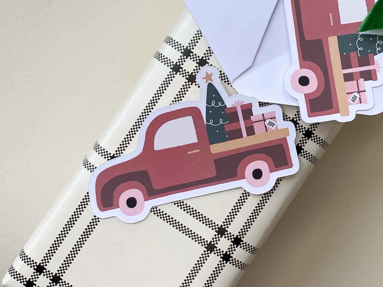 A burnt orange vintage style Christmas truck note card with festive gift boxes and a Christmas tree, resting on black-and-white plaid wrapping paper. A white envelope is partially visible in the background.