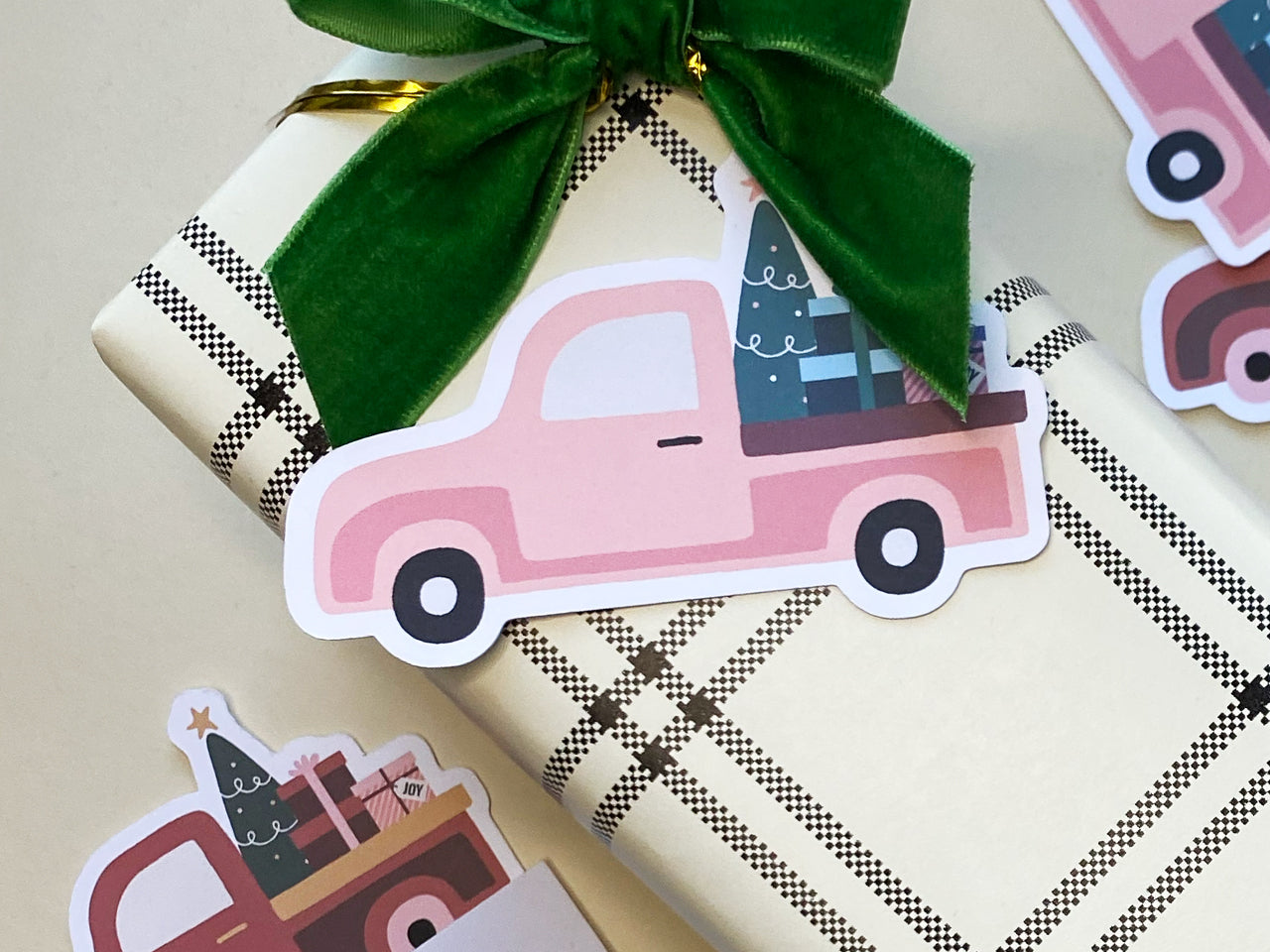 A pink vintage style Christmas truck die-cut note card placed on a plaid-wrapped gift with a large green velvet bow. The retro style truck carries a festive Christmas tree and colorful gift boxes.