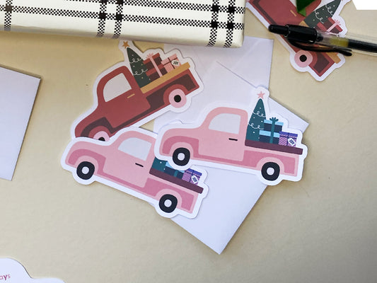 Three vintage Christmas truck die-cut mini note cards in burnt orange and pink. Each truck carries a festive load of a Christmas tree and gift boxes. The note cards are handmade, placed on a light background, with white envelopes.