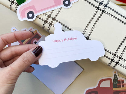 A hand with dark nail polish holding the back of a vintage style Christmas truck note card that reads Happy Holidays with lines for writing. In the background, a pink truck note card rests on plaid wrapping paper surrounded by holiday decorations.