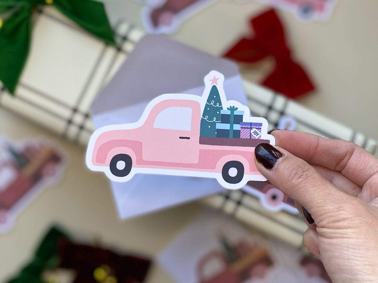 A close-up of a pink vintage Christmas truck die-cut note card being held by a human hand. The truck design features a green Christmas tree with a star on top and gift boxes in the truck bed. In the background, white envelope and a red bow.