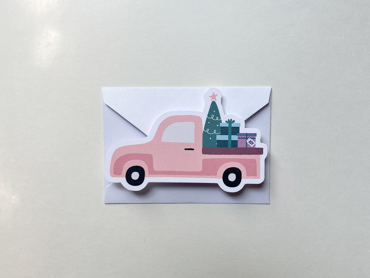 A pink vintage style Christmas truck die-cut note card placed on top of a white envelope. The card design features a Christmas tree and gift boxes in the truck bed, set against a neutral background.