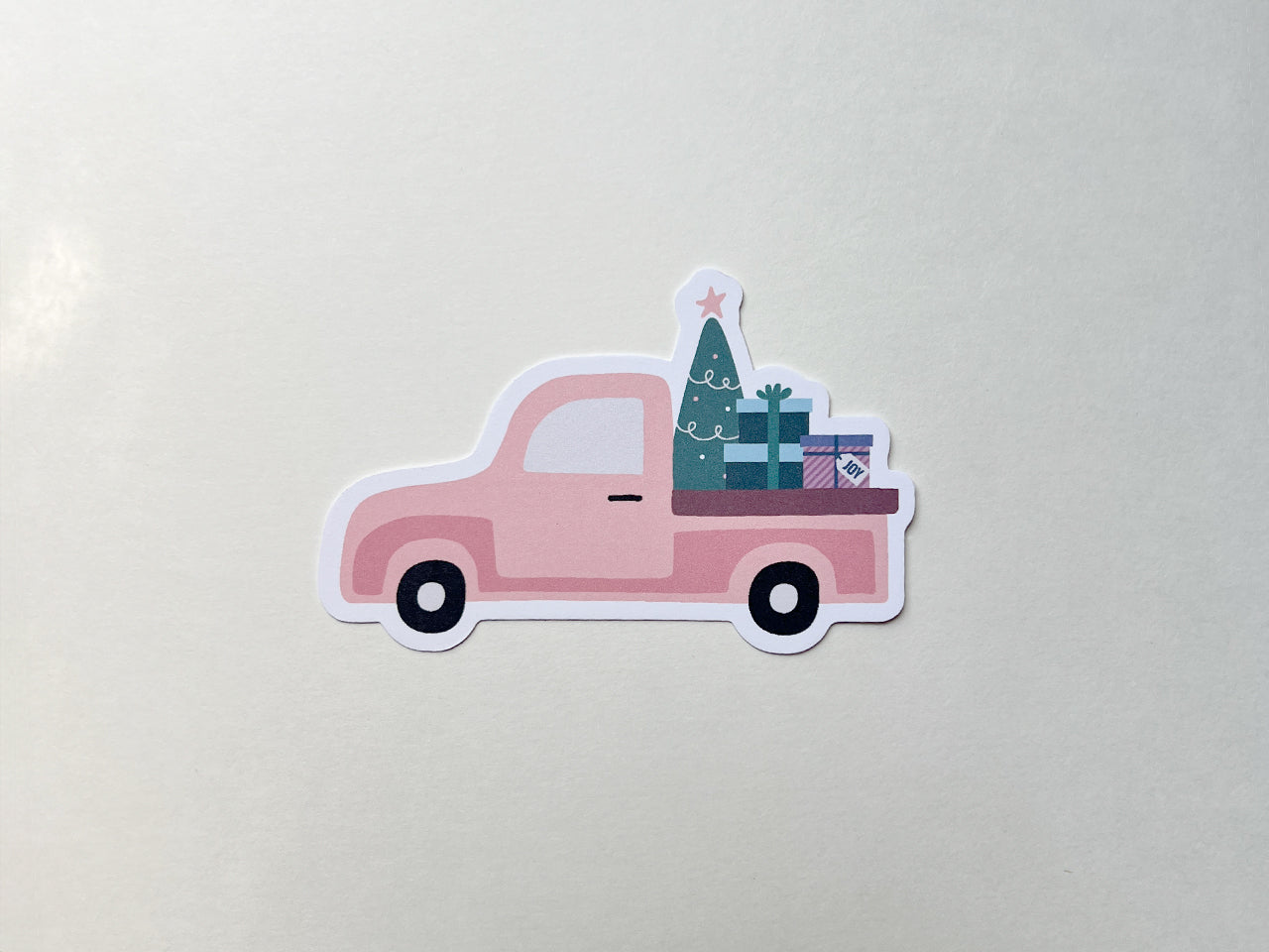 A pink vintage style Christmas truck die-cut note card featuring a festive design with a Christmas tree topped with a star and stacked gift boxes in the truck bed. The card is displayed on a plain light background.