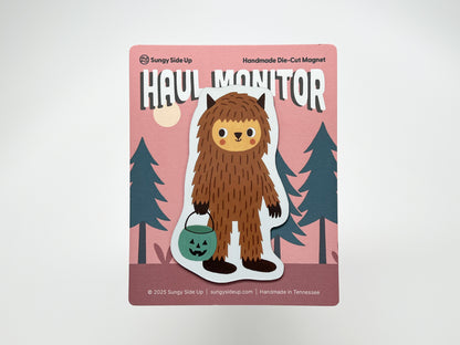 Handmade die-cut Bigfoot Halloween magnet on illustrated backing card with a pink forest background. Features a cute brown sasquatch holding a teal jack-o’-lantern candy bucket. Perfect quirky fridge décor for cryptid fans, spooky season gifts, and woodland creature collectors.