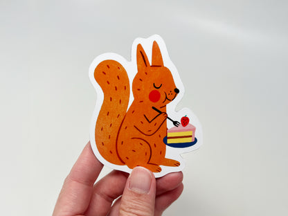 Hand holding a handmade die-cut fridge magnet shaped like an orange squirrel enjoying a slice of strawberry-topped cake. The cute woodland animal design is vibrant and playful, making it ideal for nature lovers, dessert fans, and quirky kitchen or home decor.