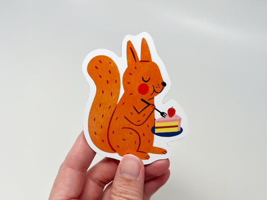 Hand holding a handmade die-cut fridge magnet shaped like an orange squirrel enjoying a slice of strawberry-topped cake. The cute woodland animal design is vibrant and playful, making it ideal for nature lovers, dessert fans, and quirky kitchen or home decor.