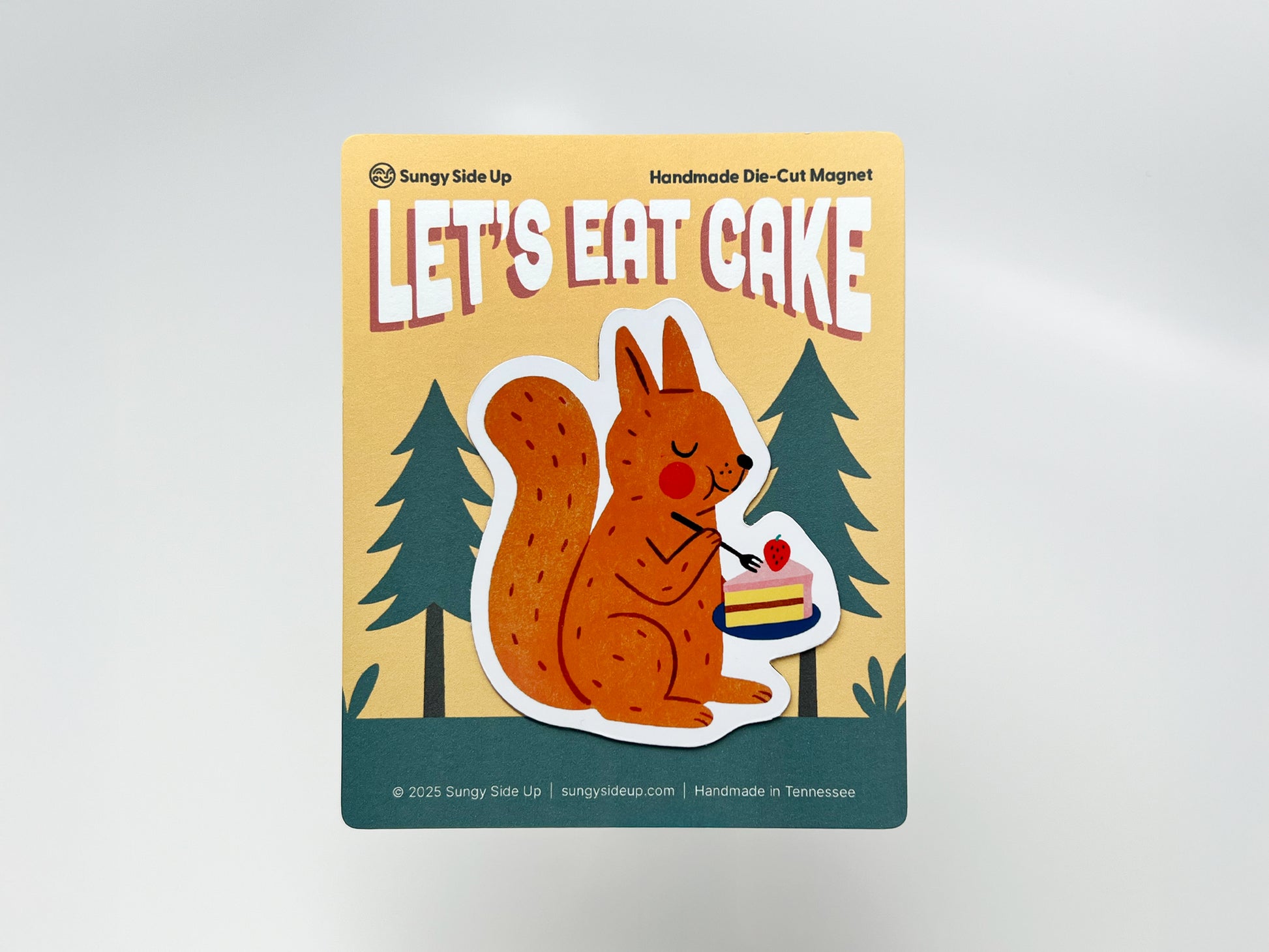 Handmade die-cut fridge magnet featuring an illustrated orange squirrel holding a plate with a slice of strawberry-topped cake, mounted on decorative backing with a woodland tree design. Perfect for animal lovers, dessert fans, and quirky kitchen or cabin decor.