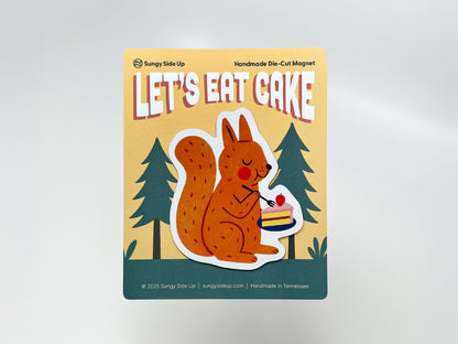 Handmade die-cut fridge magnet featuring an illustrated orange squirrel holding a plate with a slice of strawberry-topped cake, mounted on decorative backing with a woodland tree design. Perfect for animal lovers, dessert fans, and quirky kitchen or cabin decor.
