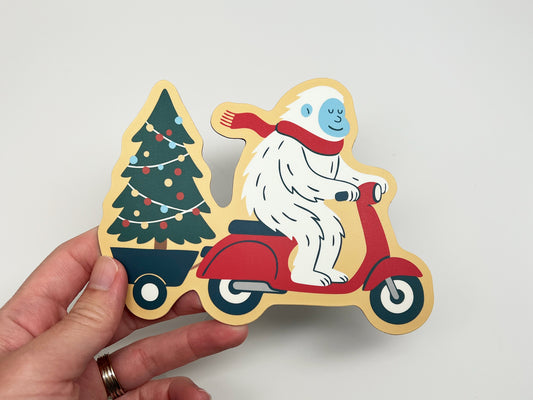 Close-up of a die-cut magnet featuring a smiling yeti riding a red scooter and towing a Christmas tree wrapped in colorful string lights. The festive illustration shows the yeti in a red scarf against a light background, emphasizing the detailed design of this holiday magnet.