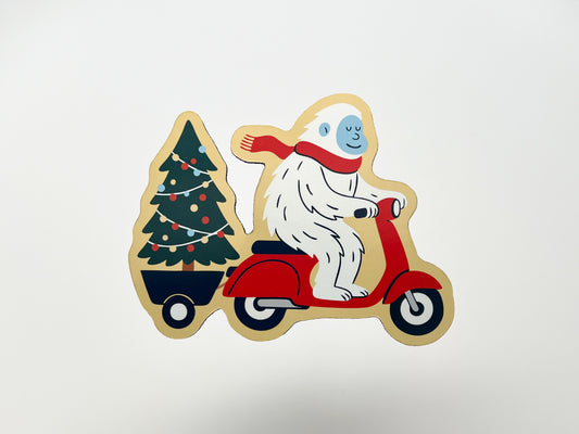 Handmade die-cut magnet of a happy yeti in a red scarf riding a red scooter and towing a decorated Christmas tree. The whimsical holiday magnet has bold lines, festive colors, and a playful design—perfect for decorating a fridge, locker, or workspace and gifting during the holiday season.