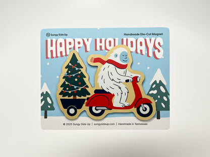 Handmade die-cut holiday magnet featuring a cheerful yeti riding a bright red scooter, hauling a decorated Christmas tree on a snowy background. The festive backing card says “Happy Holidays” in bold red letters. A quirky and fun Christmas decoration, perfect for gifting or holiday décor.
