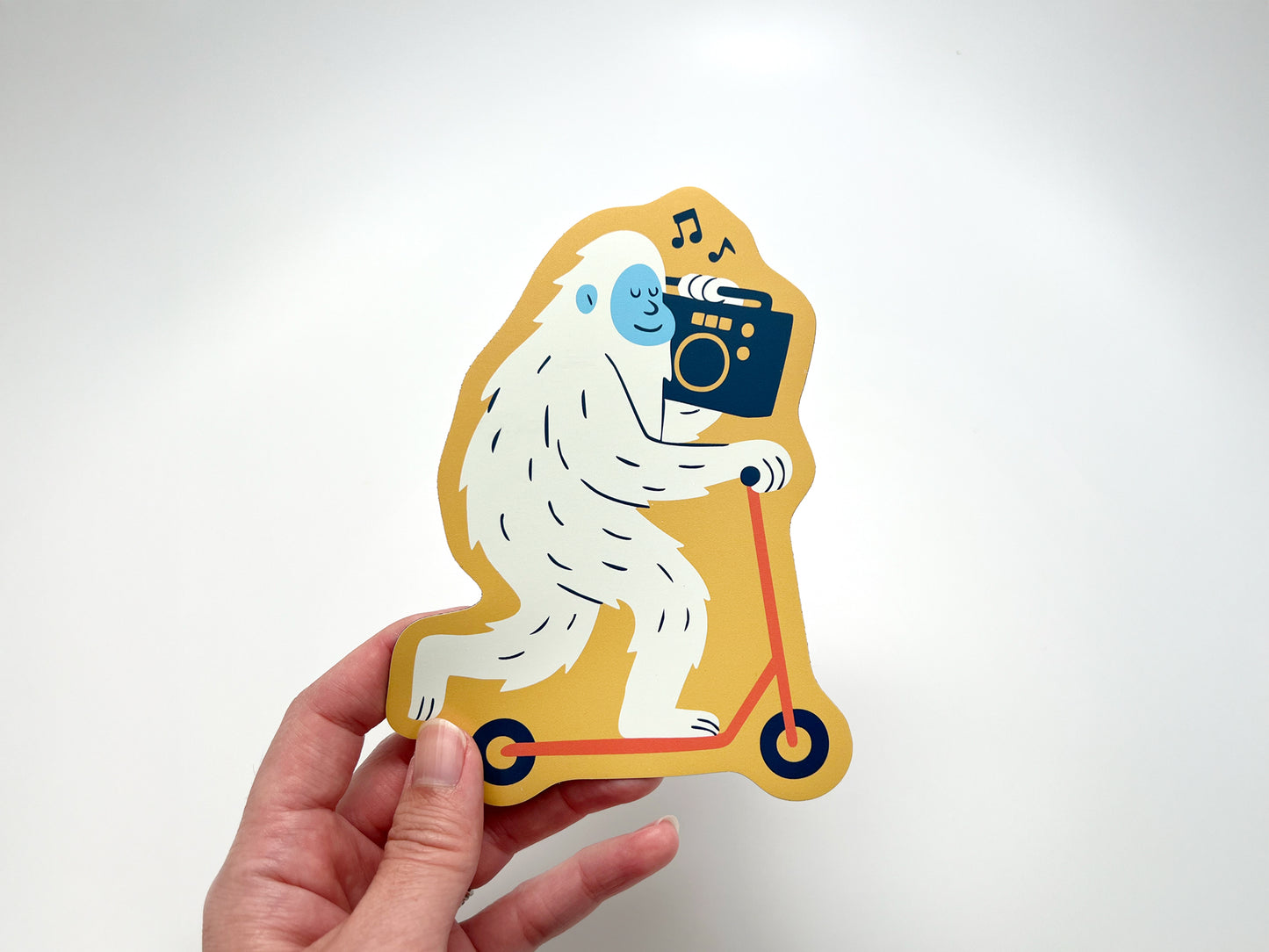 Hand holding a die-cut fridge magnet of a smiling yeti riding a yellow scooter and blasting music from a boombox. Funny and quirky novelty magnet gift for music lovers, scooter fans, and playful kitchen decor.