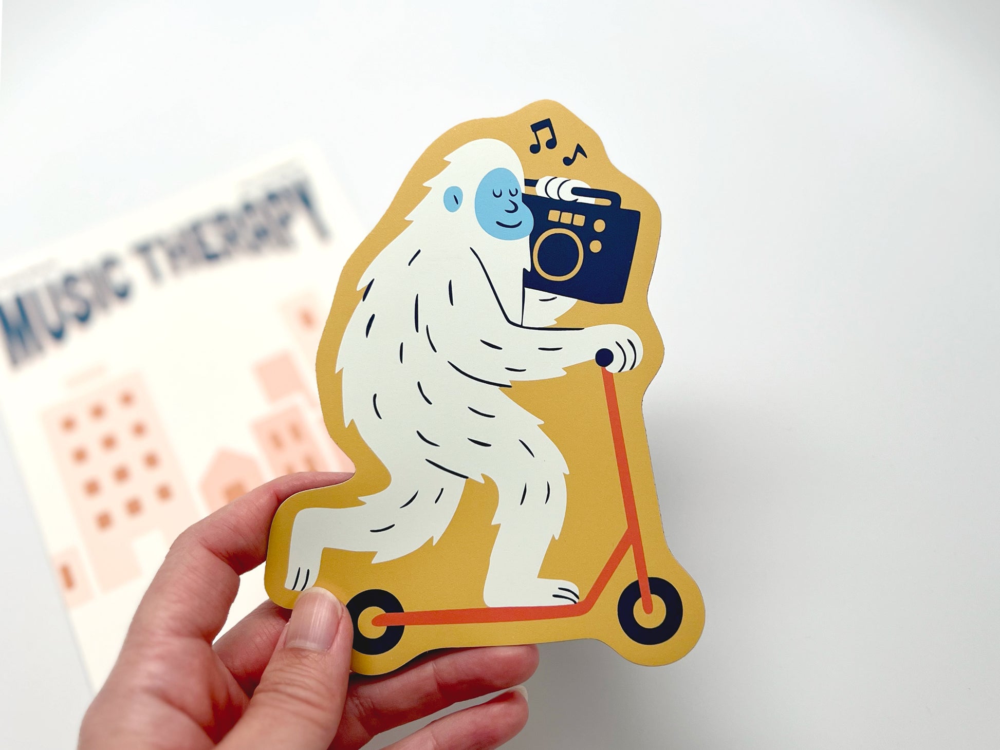 Die-cut fridge magnet featuring a quirky yeti riding a yellow scooter while carrying a black boombox with music notes. Backing card reads “Music Therapy.” Fun novelty gift for music lovers, scooter fans, and unique home decor.