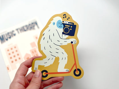 Die-cut fridge magnet featuring a quirky yeti riding a yellow scooter while carrying a black boombox with music notes. Backing card reads “Music Therapy.” Fun novelty gift for music lovers, scooter fans, and unique home decor.