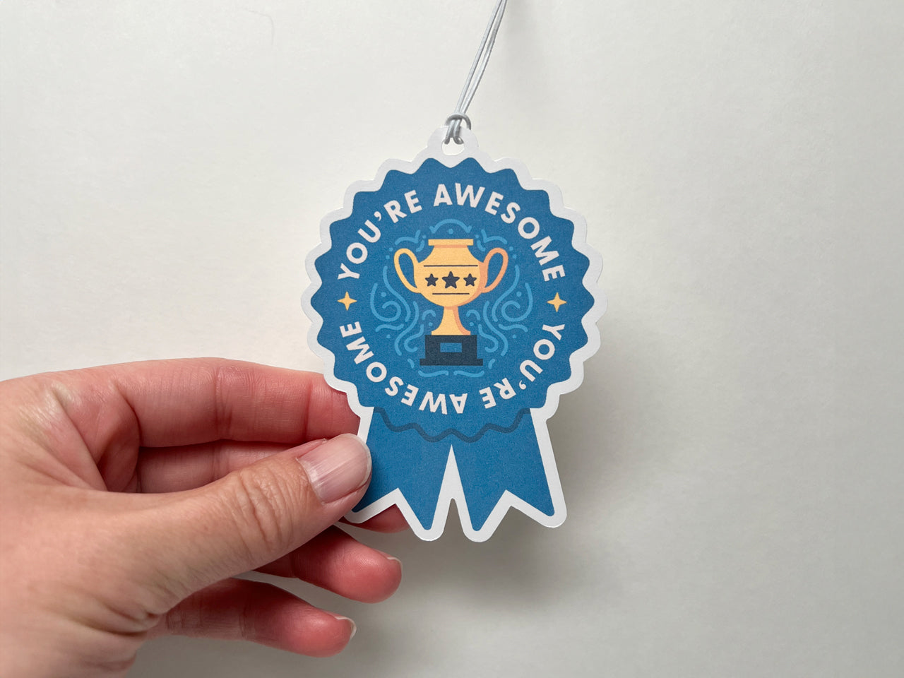 A close-up of a hand holding a blue ribbon-shaped gift tag that says “You’re Awesome” around a golden trophy illustration. The tag is attached to a white string and set against a plain white background, highlighting its colorful design and celebratory theme.