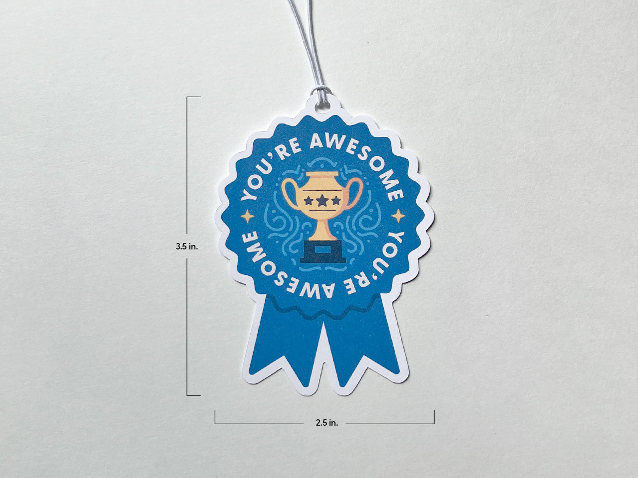 A blue ribbon-shaped gift tag with the words You're Awesome surrounding a golden trophy illustration. The gift tag measures 3.5 inches in height and 2.5 inches in width. A white string is attached at the top for easy hanging.