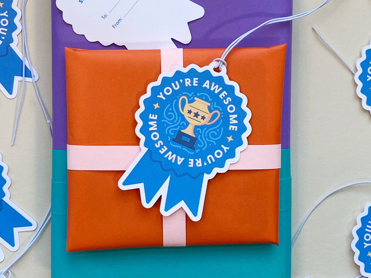 A colorful wrapped gift with a blue ribbon-shaped gift tag with the words You’re Awesome surrounding a golden trophy illustration. The scene is bright, cheerful, and celebratory, showcasing the gift tags as part of festive packaging.