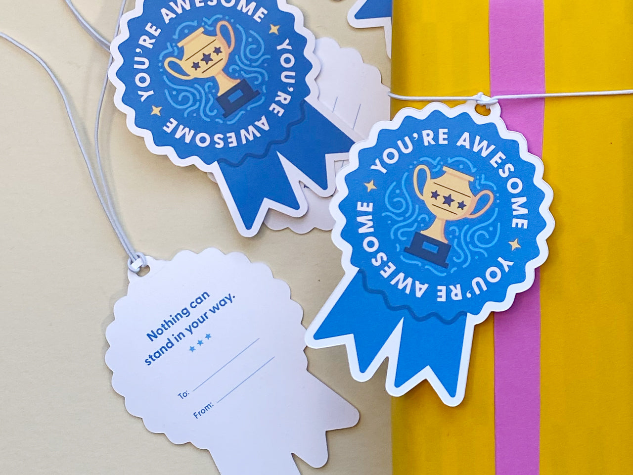 A close-up of blue ribbon-shaped gift tags with You’re Awesome text and a golden trophy illustration. One tag is flipped to show the back, featuring the text Nothing can stand in your way with space for To and From details.