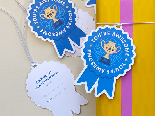 A close-up of blue ribbon-shaped gift tags with You’re Awesome text and a golden trophy illustration. One tag is flipped to show the back, featuring the text Nothing can stand in your way with space for To and From details.