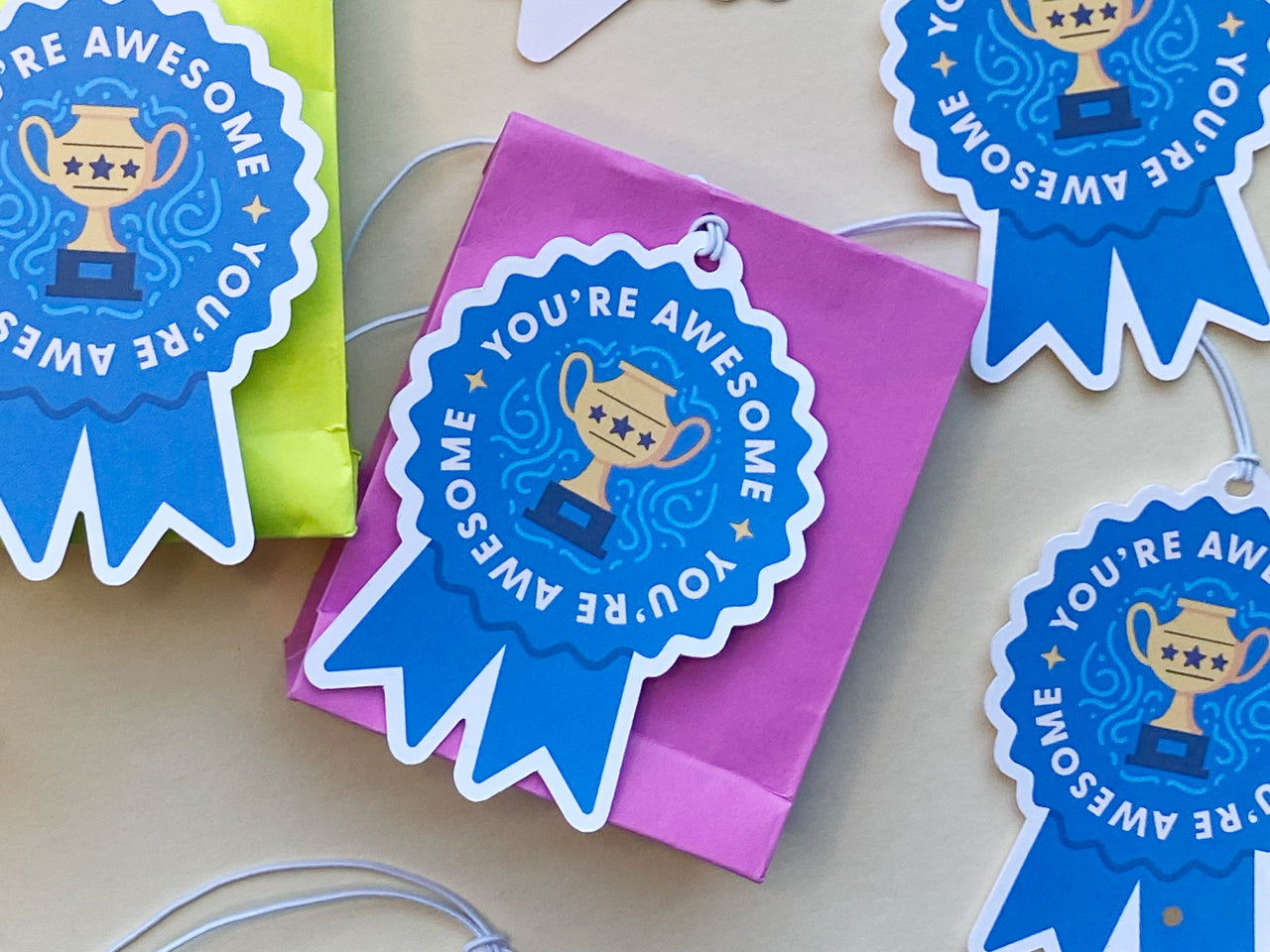 A vibrant pink gift bag features a blue ribbon-shaped tag that says You're Awesome with a golden trophy illustration. A similar tag hangs on a neon green gift bag to the side, creating a colorful and celebratory atmosphere.