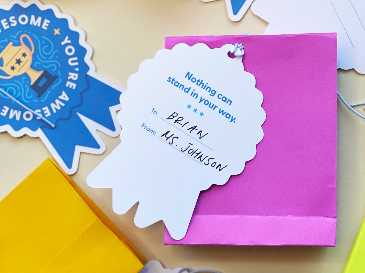 The back of a blue ribbon-shaped tag is shown, tied to a bright pink gift bag. The tag reads Nothing can stand in your way with To and From spaces. Surrounding the scene are more tags, suggesting a cheerful, personalized gift setup.