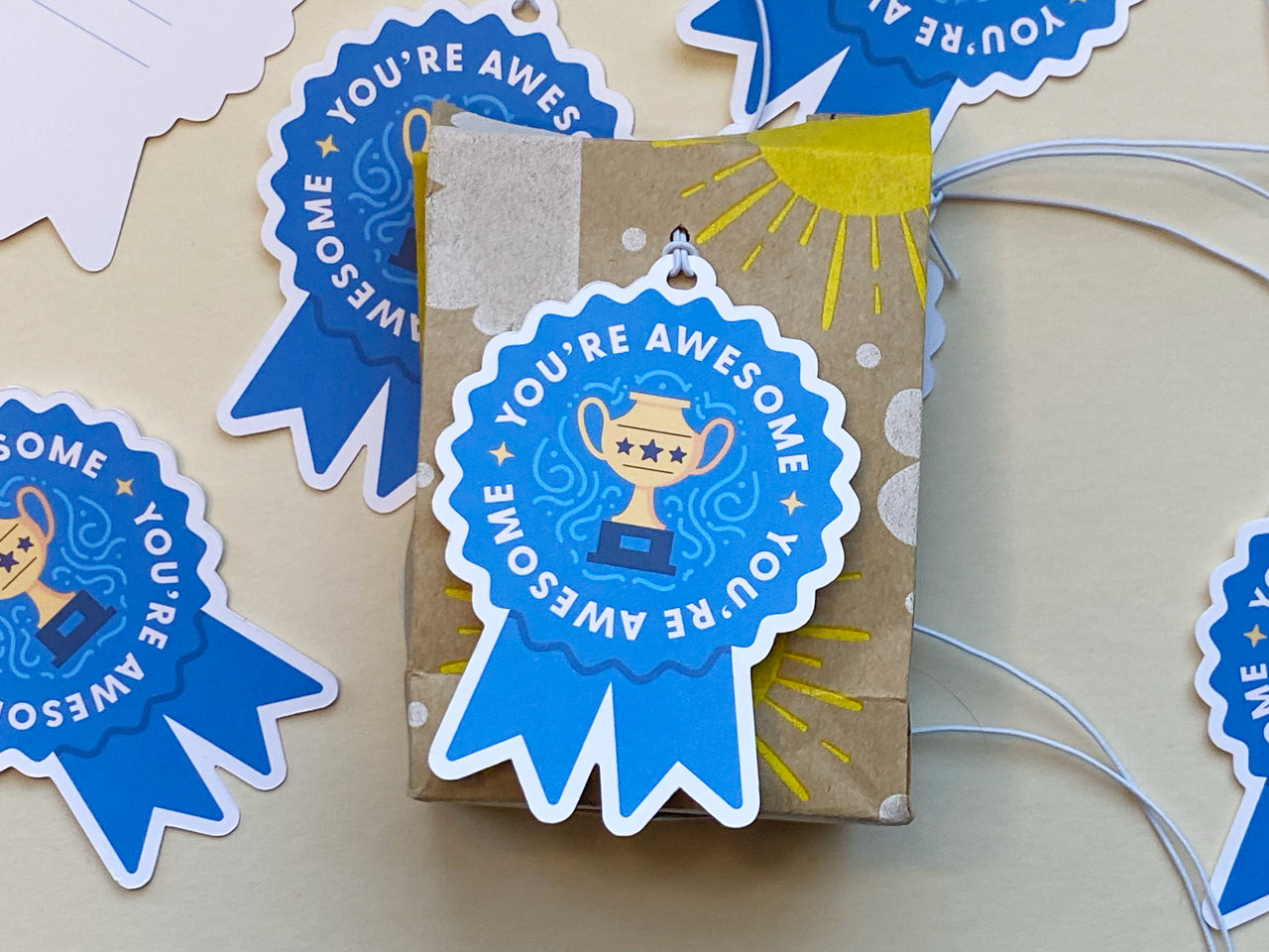 A small, brown kraft gift box decorated with hand-drawn yellow sun and dot designs is topped with a blue ribbon-shaped gift tag with white elastic string that says You’re Awesome and features a golden trophy illustration.